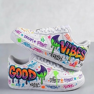 Op de afbeelding: Witte sneakers met kleurrijke graffiti-kunst. De schoenen tonen de woorden "GOOD" en "VIBES" in druipende verf, samen met "DREAM", "STREET" en "FRESH". Het ontwerp bevat sterren en verfspatten.