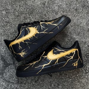 May include: Black sneakers with gold lightning bolt design. The shoes feature a gold Nike swoosh and the word "AIR" in gold near the heel. The sneakers are on a gray surface.