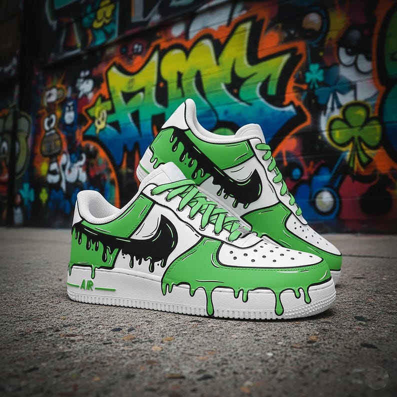 Puede incluir: Un par de zapatillas blancas con un dise&ntilde;o de pintura goteante verde y negro. Los zapatos tienen un logotipo de Nike negro y cordones verdes. La palabra "AIR" est&aacute; impresa en el lateral de la suela blanca. El fondo presenta arte de graffiti colorido.
