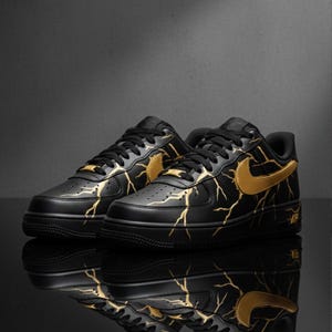 May include: Pair of black sneakers with gold lightning bolt designs and gold swoosh logos. The shoes have black laces and soles. The word "AIR" is visible on the side of the shoe.