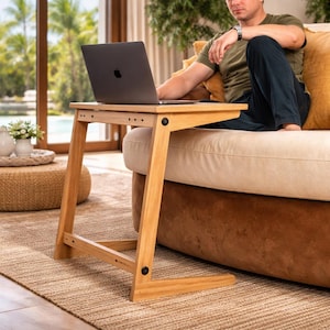 May include: A light-colored wooden laptop table with a rectangular top and angled legs. The table is positioned next to a sofa, with a laptop on top. The table is made of light-colored wood and has a simple, modern design.