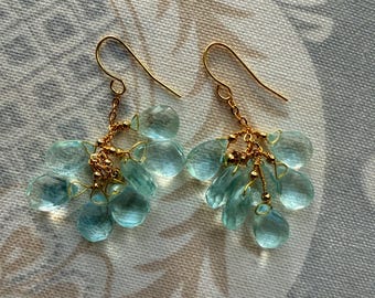 May include: A pair of gold-tone earrings featuring clusters of light blue, teardrop-shaped gemstones. The earrings have a delicate, dangling design with a hook closure. The gemstones are faceted, catching the light.