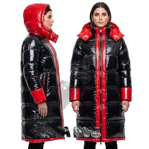 Peut inclure: Un long manteau doudoune noir brillant avec une capuche rouge vif, des poignets rouges et une patte de fermeture éclair verticale rouge. La veste matelassée possède un col haut et une finition brillante, vue de profil et de face.