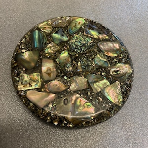 May include: A round, decorative coaster with a mosaic design. The coaster features iridescent abalone shells and small, dark-colored stones set in a clear resin. The shells have shades of green, blue, and gold.
