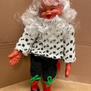 Puede incluir: Una muñeca vintage de Papá Noel con una cara sonriente, barba blanca y gorro rojo. La muñeca viste un suéter blanco con lunares negros, pantalones negros y botas rojas con zapatos negros. Lazos verdes adornan las botas.