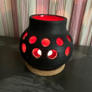 Könnte beinhalten: Ein schwarzer Keramik-Kerzenhalter mit roter Innenseite und kreisförmigen Aussparungen. Der Kerzenhalter steht auf einem Korksockel. Das Innere und die Aussparungen leuchten durch das Licht einer Kerze.