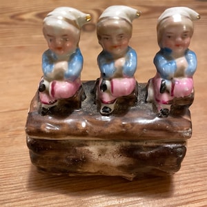 May include: A vintage ceramic trinket box featuring three seated figures. Each figure wears a white hat with gold trim, a blue jacket, and pink pants. The figures sit on a brown, log-shaped base, atop a rectangular box.