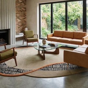 Può includere: Un set da soggiorno con due divani in pelle beige con gambe dorate, due poltrone verde oliva con struttura in legno e un tavolino con piano in vetro. Sotto è posizionato un grande tappeto circolare beige con un bordo grafico bianco e nero.