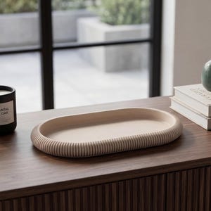 Op de afbeelding: Een houten ovaal dienblad met een geribbelde rand op een donker houten oppervlak, naast een zwarte kaars met het label "Santal & Oak". Een kleine groene vaas met gedroogd gras en een vetplant staan op een stapel boeken.