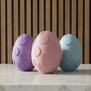 May include: Three decorative Easter eggs in pastel colors: lavender, pink, and light blue. Each egg features a pattern of raised, circular dots. The eggs are displayed on a marble surface against a backdrop of vertical wooden slats.