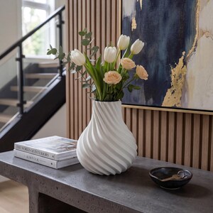 May include: A white, swirled vase holds white tulips and peach roses. The vase sits on a gray concrete console table, next to a stack of books and a small black bowl. A painting with gold accents hangs in the background.