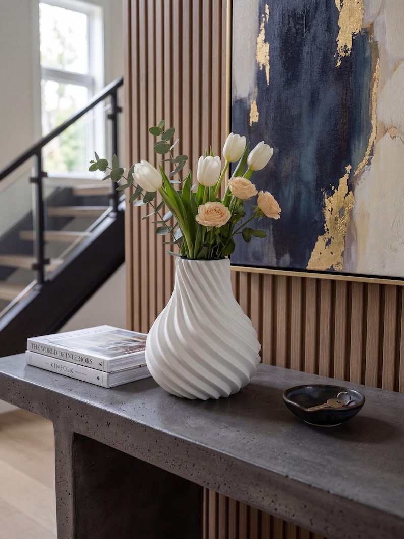 May include: A white, swirled vase holds white tulips and peach roses. The vase sits on a gray concrete console table, next to a stack of books and a small black bowl. A painting with gold accents hangs in the background.