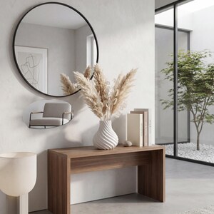 May include: A modern interior scene featuring a wooden console table with a white vase holding dried pampas grass. A large round black-framed mirror hangs above the table, reflecting a small chair. Two books and a decorative stone are on the table.
