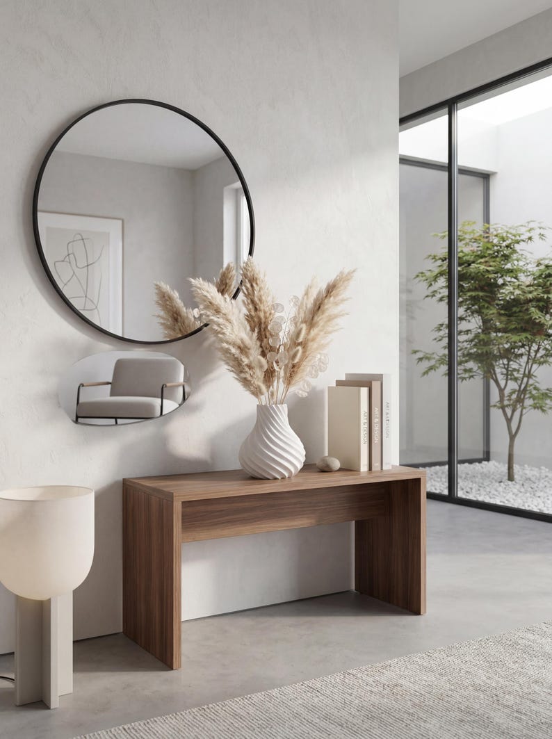 May include: A modern interior scene featuring a wooden console table with a white vase holding dried pampas grass. A large round black-framed mirror hangs above the table, reflecting a small chair. Two books and a decorative stone are on the table.