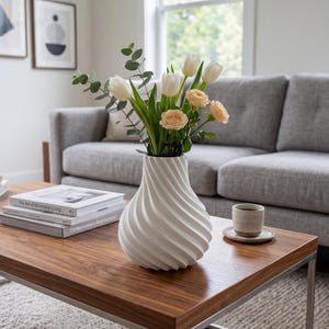 May include: A white, spiral-textured vase holding white tulips and peach roses. The vase sits on a dark wood coffee table with a metal frame. Books and a cup of coffee are also on the table.