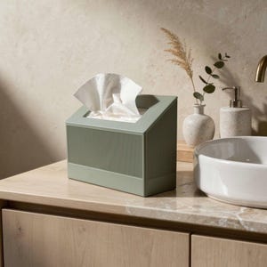 May include: A sage green rectangular tissue box cover with a slanted top and vertical ribbed texture on the sides. The box is constructed from a solid material and features a wide rectangular opening at the top, through which white tissues emerge.