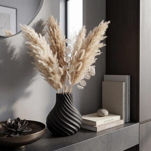 May include: A black, spiral-patterned vase holds dried pampas grass and delicate, round, silver-dollar eucalyptus. A bronze-toned bowl with a succulent sits to the left. Books and a stone accent the shelf.
