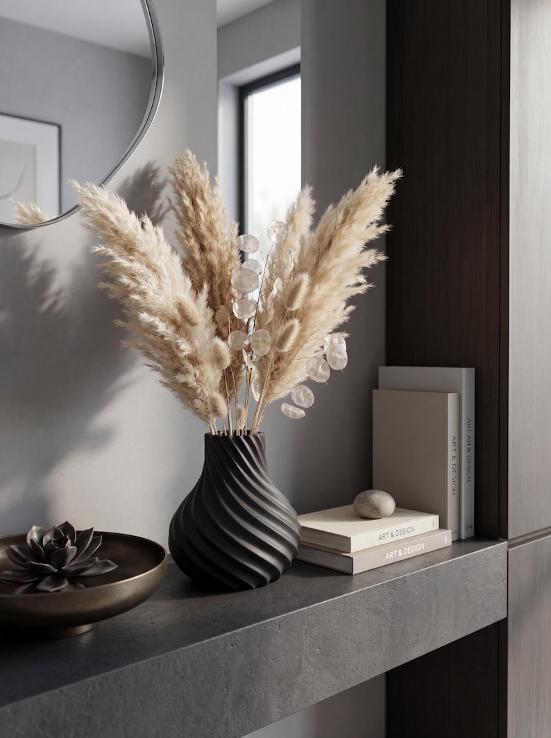 May include: A black, spiral-patterned vase holds dried pampas grass and delicate, round, silver-dollar eucalyptus. A bronze-toned bowl with a succulent sits to the left. Books and a stone accent the shelf.