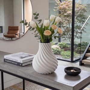 White twisted spiral ribbed vase with fresh tulips and eucalyptus – modern 3D printed PLA swirling diagonal texture. Minimalist Scandinavian Japandi home decor on concrete console with mirror and fresh flowers