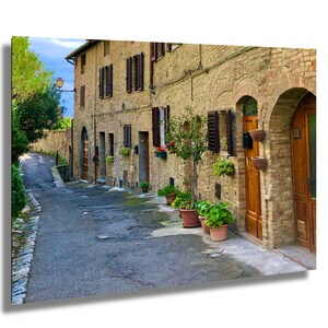 Puede incluir: Imagen de una calle estrecha adoquinada en un antiguo pueblo europeo. Los edificios con muros de piedra, contraventanas de madera y puertas arqueadas bordean la calle. Las plantas en macetas y los árboles añaden vegetación. El cielo es azul.