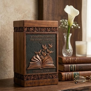 Peut inclure: Une urne funéraire rectangulaire en bois avec un panneau central sombre orné d'un relief représentant un livre ouvert et des oiseaux en vol. Le texte indique A CHAPTER CLOSED, THE STORY LIVES ON, ETERNAL LOVE et EMBRACED BY MEMORIES en relief.