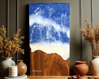 May include: A rectangular wooden artwork showing a beach scene. The top section has a blue and white wave design, leading to a sandy area, and then a brown wooden base. The word "Ashrests" is written on the wood. The artwork is displayed on a wooden surface.