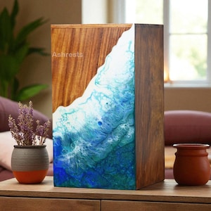 May include: A wooden box with a unique design. The top half is a natural wood grain, while the bottom half features a blue and white resin design resembling ocean waves. The word "Ashrests" is printed on the wood.