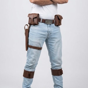 May include: Brown leather utility belt with two pouches, a holster, and leg straps. The belt has a silver buckle and is worn with light blue jeans and a white t-shirt. The leather accessories are a rich, dark brown color.