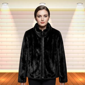 May include: A black faux fur jacket with a high collar and a full-length zipper. The jacket has long sleeves and a slightly rounded hem. The model is wearing the jacket in front of a brick wall.