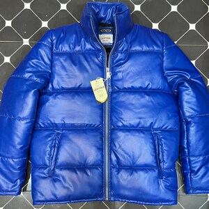Puede incluir: Chaqueta acolchada azul brillante con cuello alto y cremallera completa. La chaqueta presenta un diseño acolchado y dos bolsillos delanteros. Una etiqueta con el texto "LEATHER" está adjunta. La chaqueta es de material brillante.