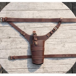 May include: A brown leather holster with a cup-shaped design, featuring a belt and shoulder straps. The holster has a buckle closure and is stitched with white thread. The straps are adjustable and designed to be worn around the waist and over the shoulder.