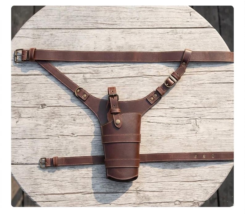 May include: A brown leather holster with a cup-shaped design, featuring a belt and shoulder straps. The holster has a buckle closure and is stitched with white thread. The straps are adjustable and designed to be worn around the waist and over the shoulder.