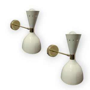 Può includere: Due applique da parete moderne della metà del secolo. Ogni applique presenta un paralume bianco a forma di clessidra con una sezione superiore grigia e dettagli in ottone. Le applique hanno un supporto a parete rotondo in ottone e un braccio in ottone.