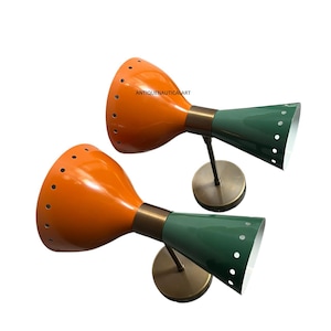 May include: Two mid-century modern wall sconces. The lamps feature orange and green conical shades with small perforations. The shades are connected by brass-colored hardware to a circular base. The text "ANTIQUENAUTICALART" is visible on the top right.