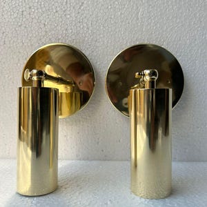 Op de afbeelding: Twee goudkleurige wandkandelaars. Elke kandelaar heeft een cirkelvormige achterplaat, een cilindrisch lichaam en een kleine, decoratieve bovenkant. De kandelaars hebben een gepolijst, reflecterend oppervlak en zijn op een witte muur gemonteerd.