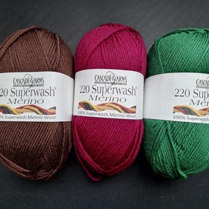 Op de afbeelding: Drie bollen Cascade Yarns 220 Superwash Merino wol in bruin, magenta en groen. Elke bol heeft een wit etiket met de merknaam en productdetails. Het garen is 100% Superwash Merino Wol.
