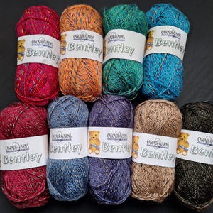 Op de afbeelding: Een verzameling van tien strengen Cascade Yarns Bentley garen in verschillende kleuren. Het garen is veelkleurig met een tweed-effect. Elke streng heeft een wit label met de merknaam en een teddybeerlogo.