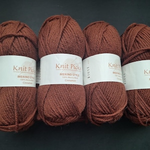 Può includere: Quattro gomitoli di filato Knit Picks Merino Style in un caldo colore cannella. Ogni gomitolo ha un'etichetta bianca con il nome del marchio e i dettagli del filato. Il filato è 100% lana merino.