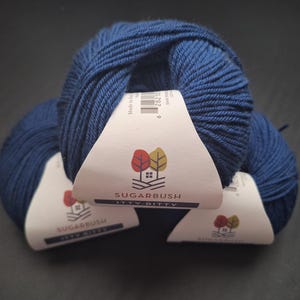 May include: Three balls of navy blue yarn with white labels featuring a logo of a house and trees. The yarn is from Sugarbush, Itty Bitty. The yarn is a deep, saturated blue, and the labels are white with a logo.