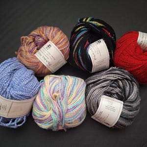 May include: A selection of yarn skeins in various colours, including red, blue, purple, and black. Each skein has a label attached. The yarn is arranged on a dark surface, displaying the range of colours and textures.