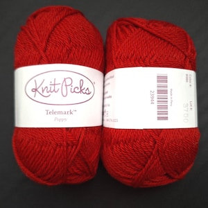 Può includere: Due gomitoli di filato rosso, etichettati "Knit Picks Telemark Poppy". Il filato è di un colore rosso vibrante, con etichette bianche che includono il nome del marchio e i dettagli del prodotto. Lo sfondo è di colore scuro e uniforme.