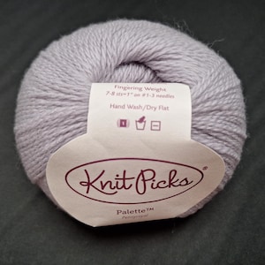 Può includere: Un gomitolo di filato lilla chiaro con un'etichetta Knit Picks. L'etichetta indica che il filato è di peso fingering e deve essere lavato a mano e asciugato in piano. Il filato è di un colore pastello.