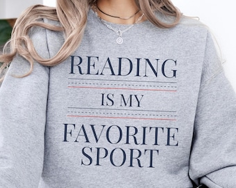 Könnte beinhalten: Ein graues Sweatshirt mit dem Text "READING IS MY FAVORITE SPORT" in Marineblau. Der Text ist mittig auf der Brust platziert, mit roten und schwarzen Linien, die die Wörter trennen. Das Sweatshirt ist ein lässiger Stil.