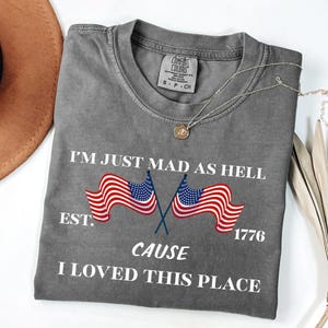 May include: A grey Comfort Colors t-shirt featuring the text "I'M JUST MAD AS HELL CAUSE I LOVED THIS PLACE" and two American flags. The shirt also has the text "EST. 1776". A silver necklace with a pendant is on the shirt.