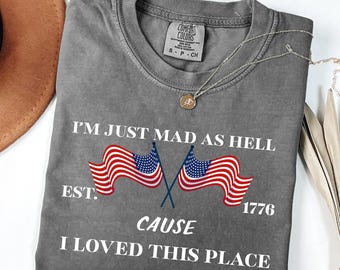 May include: A gray Comfort Colors t-shirt with the text "I'M JUST MAD AS HELL CAUSE I LOVED THIS PLACE" and two American flags. The shirt also has the text "EST. 1776". A silver necklace with a pendant is on the shirt.
