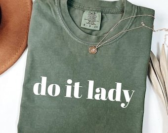 Puede incluir: Una camiseta verde oliva con la frase "do it lady" impresa en blanco. Un collar dorado con un colgante está drapeado sobre la camiseta. La camiseta está doblada sobre una superficie blanca, con un sombrero marrón visible en el fondo.