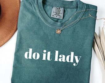 Puede incluir: Una camiseta Comfort Colors de color verde azulado con la frase "do it lady" en letras blancas. Un collar dorado con un colgante circular descansa sobre la camiseta. La camiseta está doblada sobre una superficie blanca.