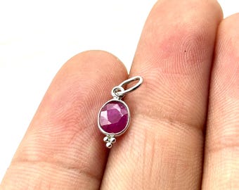 May include: A small silver pendant featuring a faceted oval-shaped deep pink ruby gemstone set in a smooth metal bezel. The pendant includes a small silver jump ring at the top and three tiny decorative metal spheres at the bottom base.