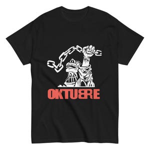 Puede incluir: Camiseta negra con un gráfico blanco de un puño levantado sosteniendo una cadena, con una cara estilizada debajo. La palabra "OKTUBRE" está impresa en letras rojas en la parte inferior de la camiseta.