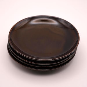 以下が含まれることがあります： 4つの濃い茶色の丸いお皿のスタック。お皿は光沢のある仕上がりで、わずかに盛り上がった縁があります。お皿は互いの上に積み重ねられており、一番上のお皿は上を向いています。